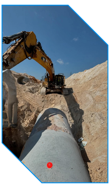 Excavator placing a large concrete pipe in a sandy trench under a clear blue sky.